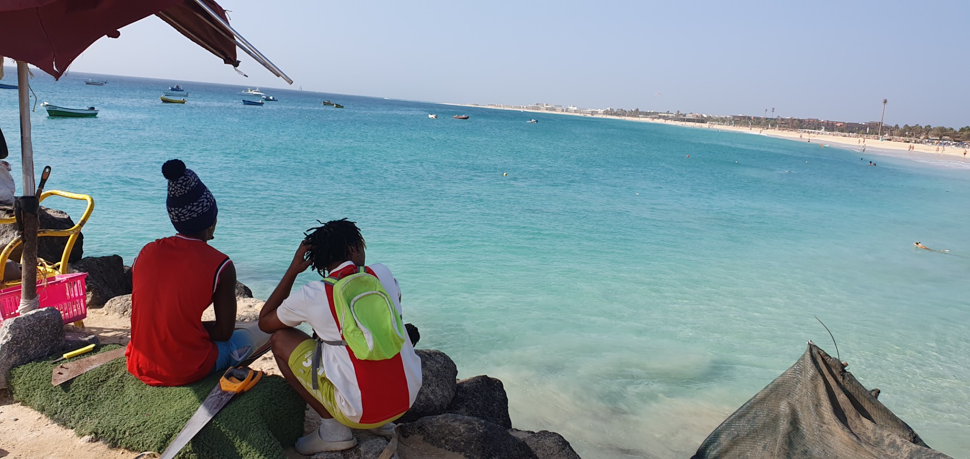 Two people sit on rocks under an umbrella, watching boats and swimmers along a sunny beach.