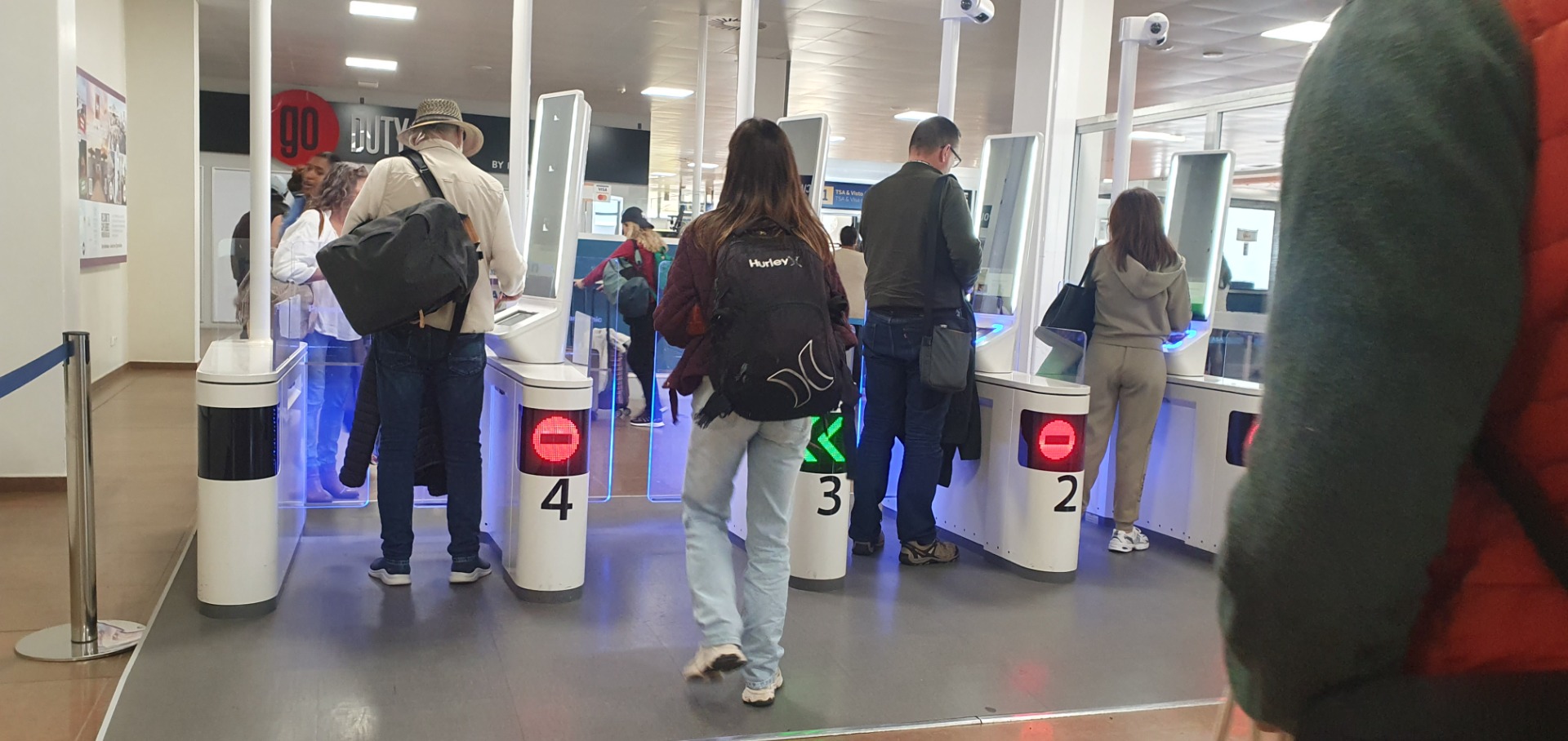 Menschen stehen vor mehreren Drehschranken mit Nummern 2 bis 4 in einem Eingangsbereich, Warteschlange.