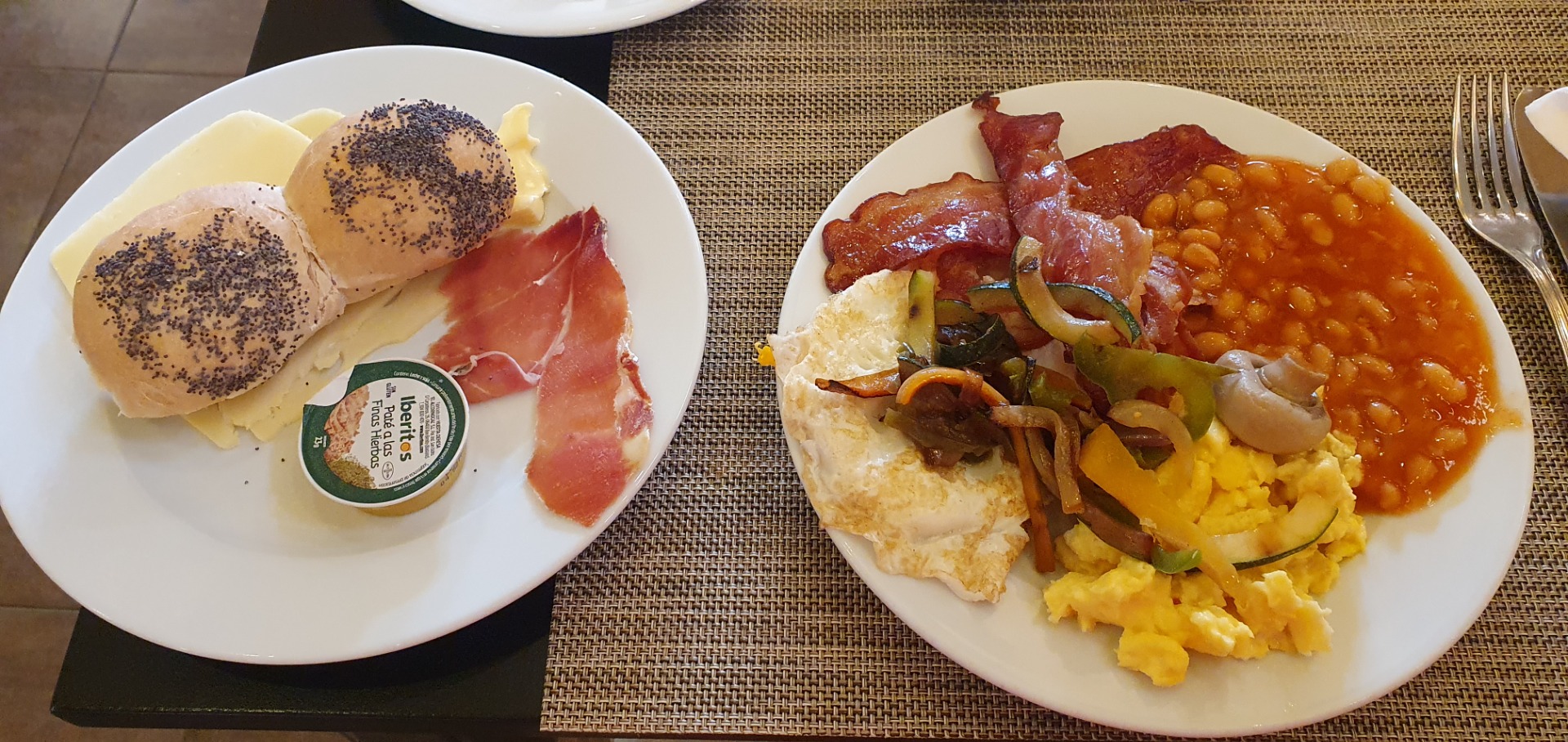 Two plates of breakfast food: poppy seed rolls with cheese and ham, plus eggs, beans, and bacon.