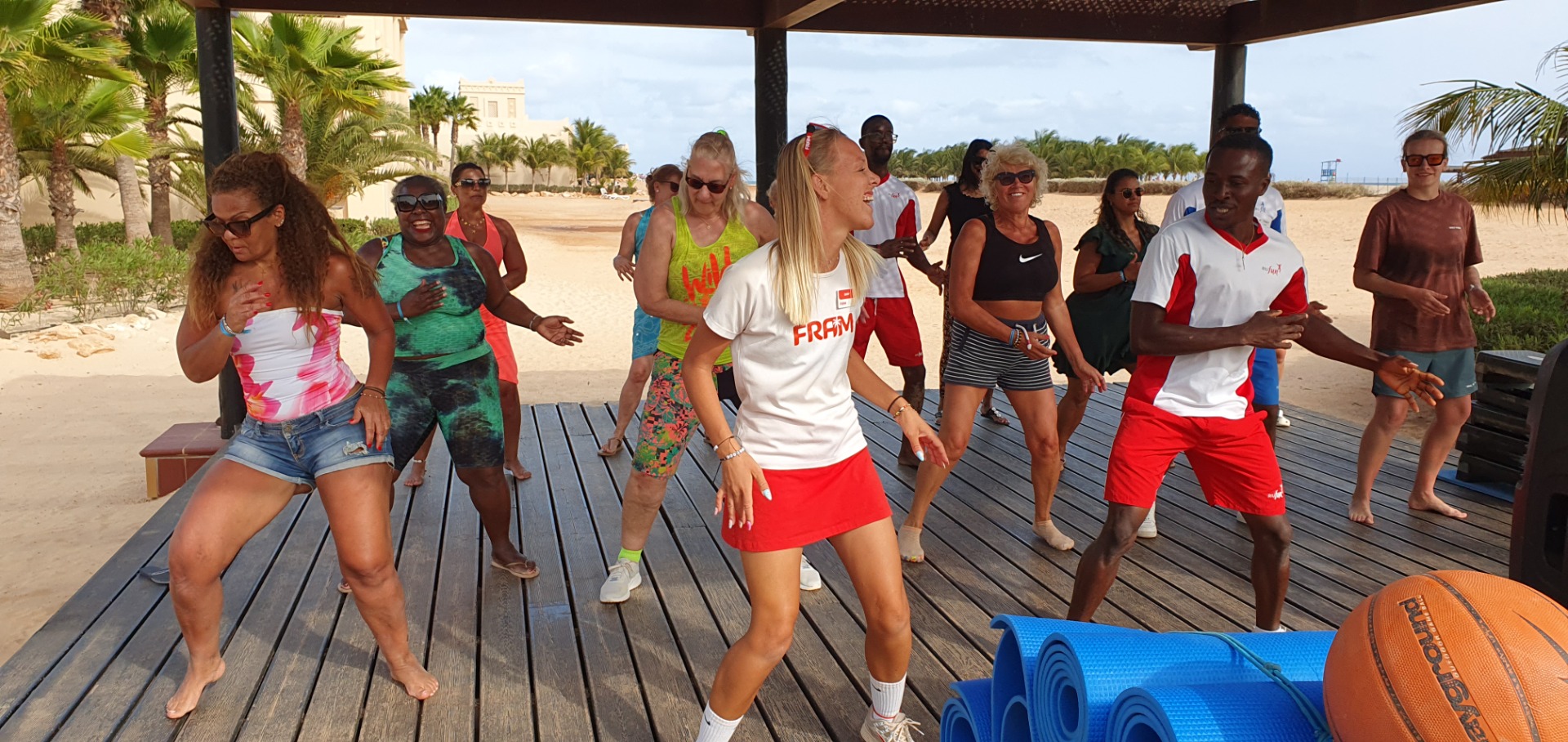 Group of people exercising on a wooden deck at a sunny beach, with yoga mats and ball.
