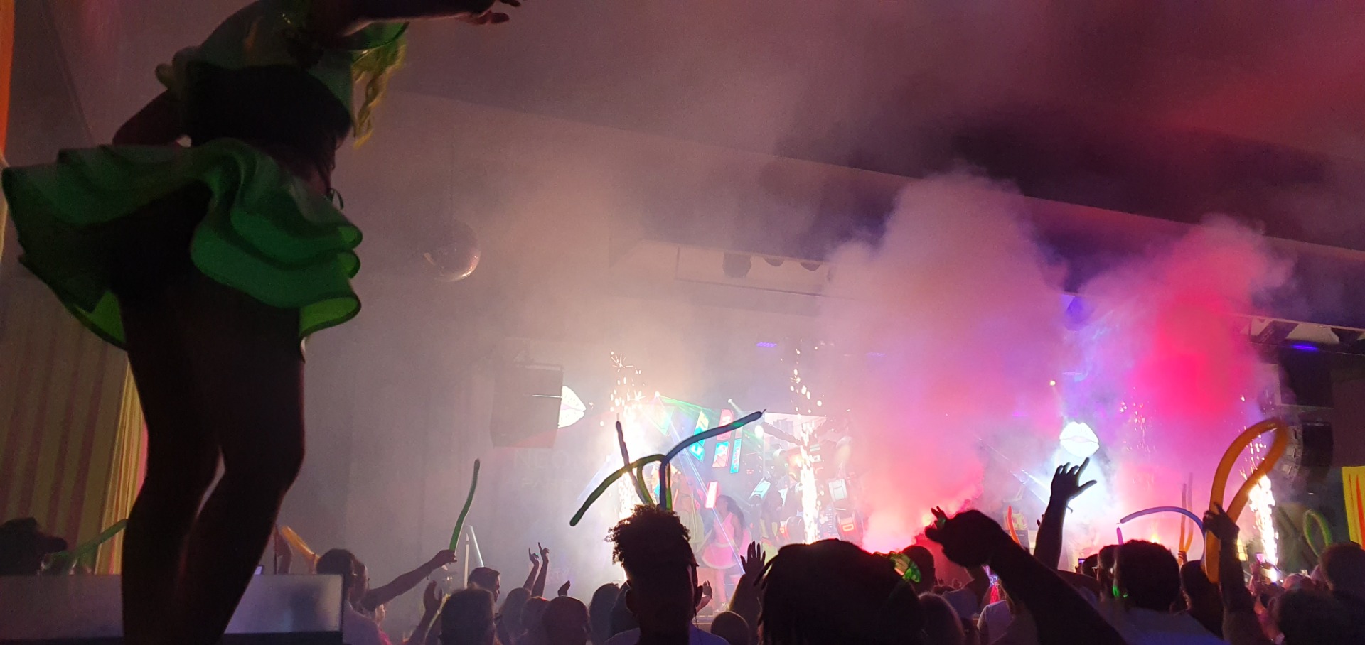 Crowd raises hands at a concert as a band performs under colorful stage lights and smoke.