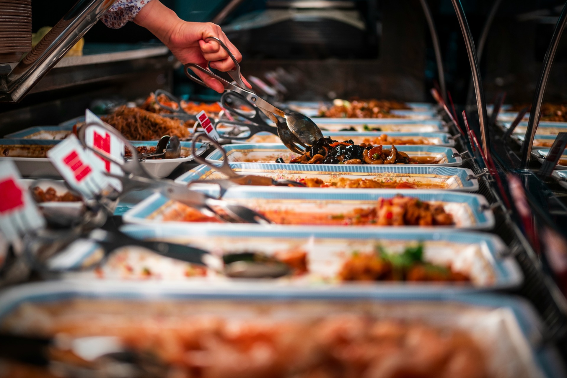 Hand holding tongs serves saucy food from buffet trays in a catering display.