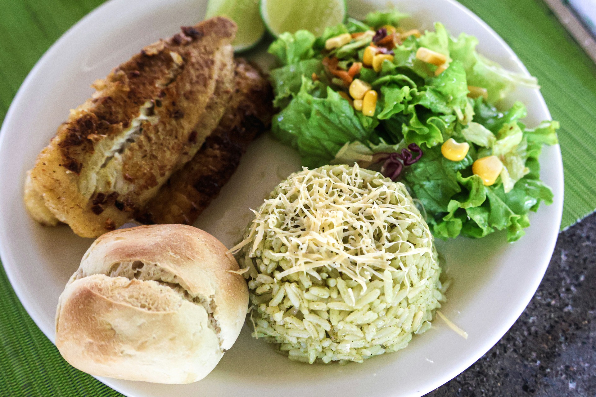 Plate with grilled chicken, mixed greens salad, corn, lime wedge, rice topped with cheese, and bread roll.