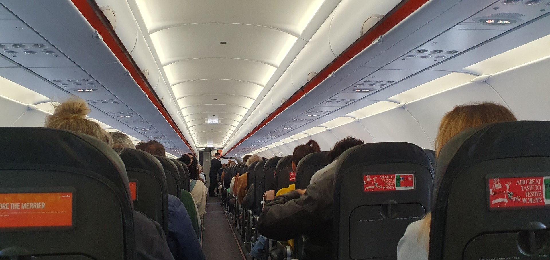 View down an airplane cabin aisle with rows of seats, passengers, and overhead lighting.