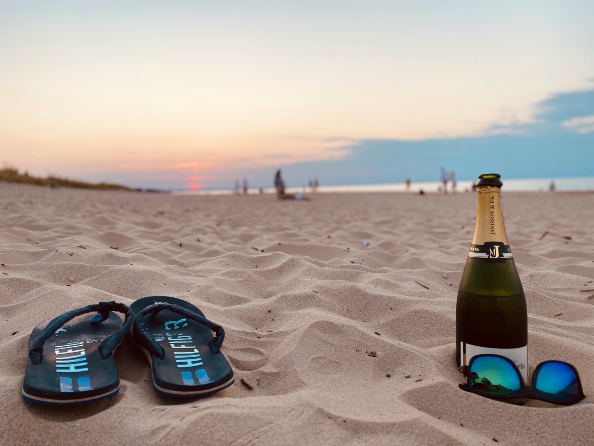 Sand beach at sunset with flip-flops and a champagne bottle in the foreground, distant people blurred.