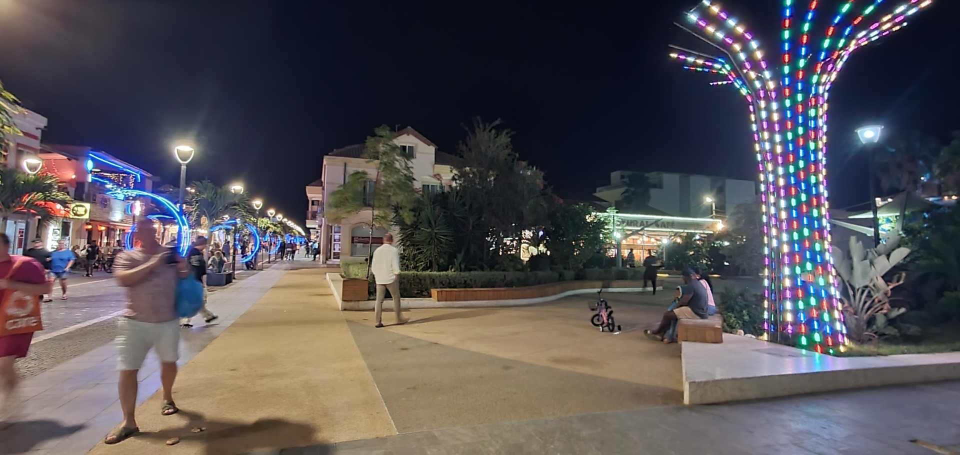 Nighttime promenade with colorful LED light columns, palm trees, and pedestrians walking past shops.