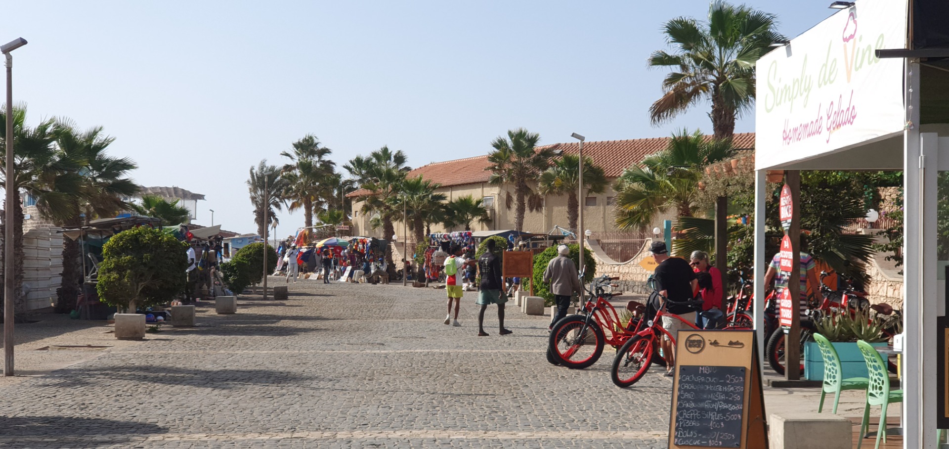 Sunny seaside promenade with palm trees, people walking, and red rental bikes near a storefront.