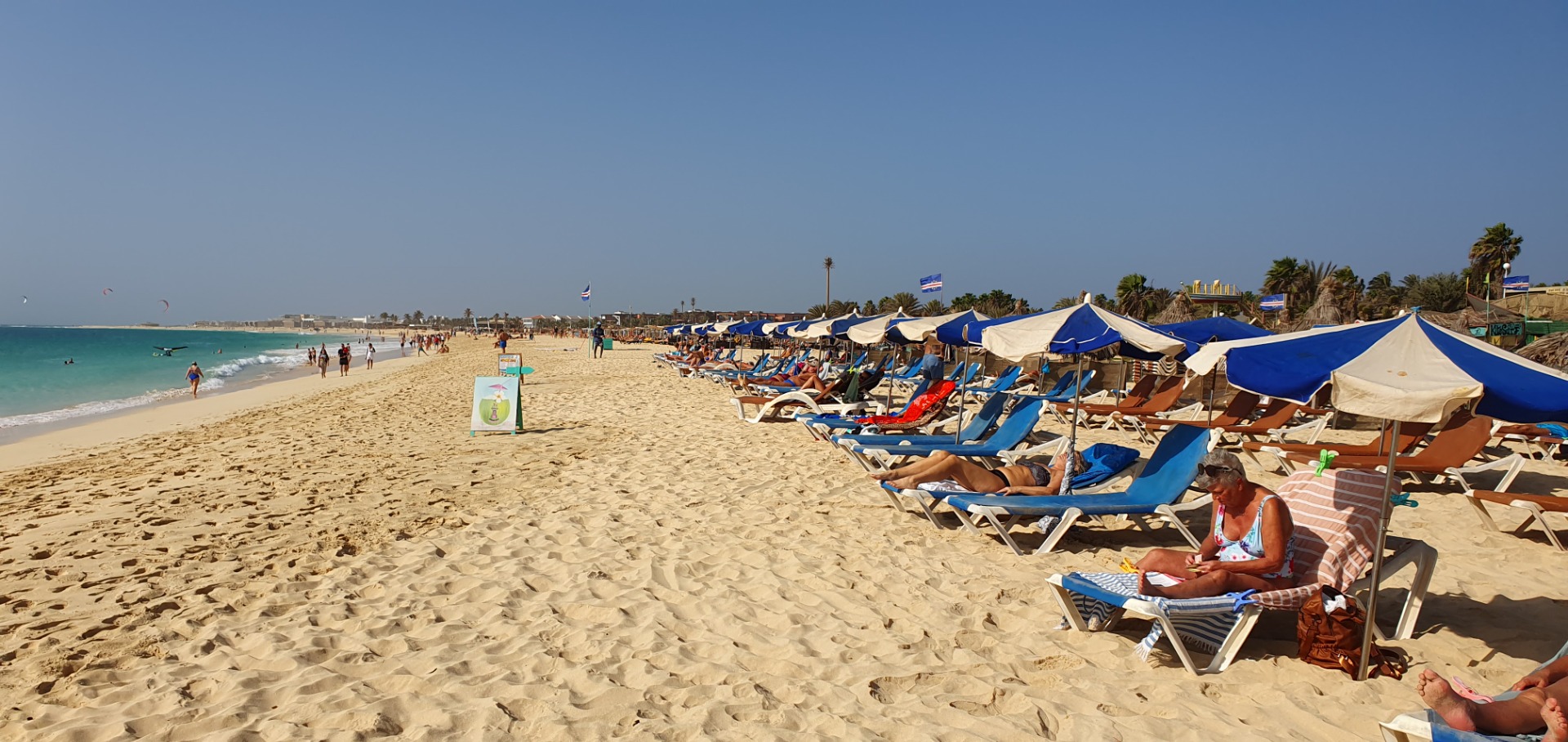 Tropischer Sandstrand mit Liegen unter Sonnenschirmen, Menschen entspannen am Meer bei klarem Himmel.