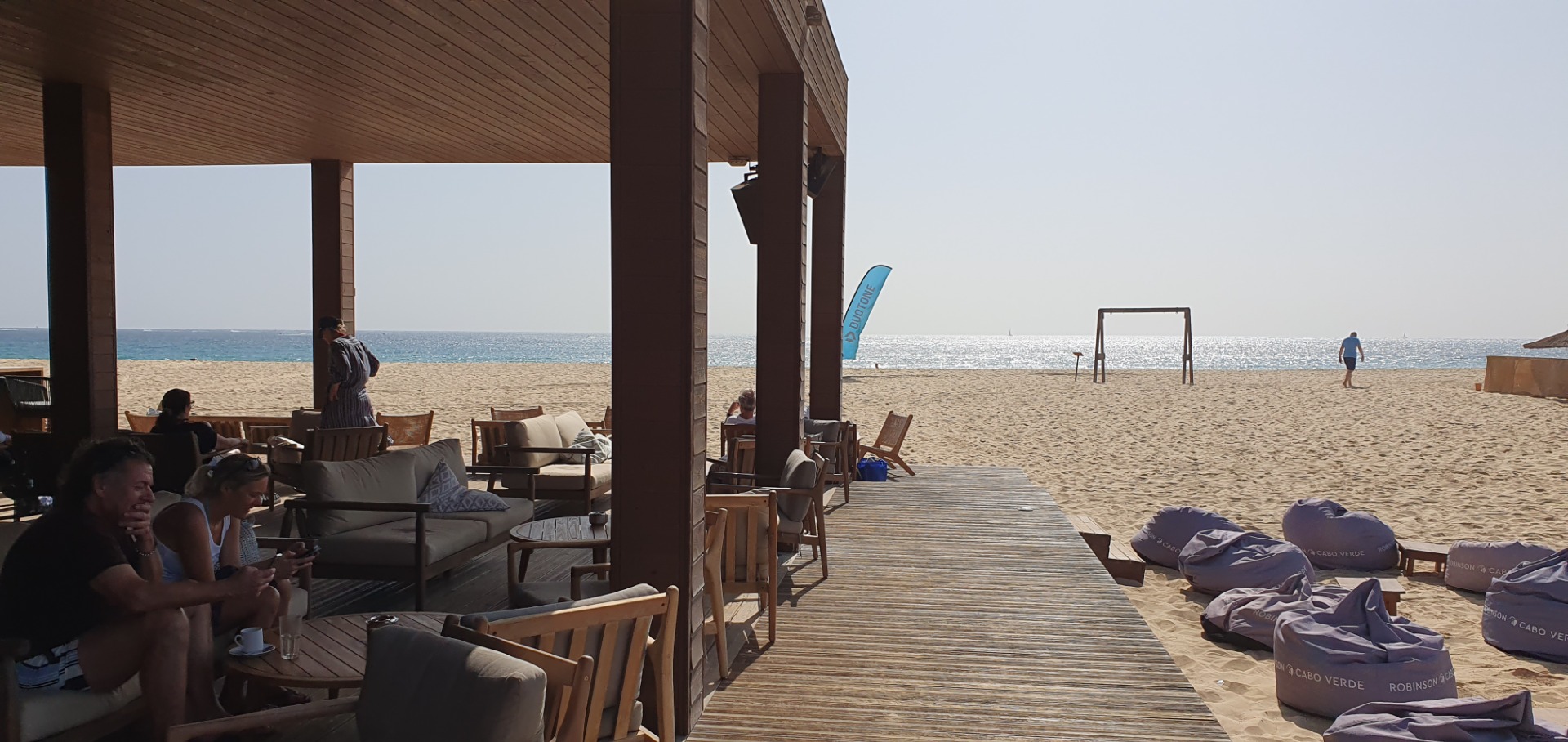 Beachside lounge with people relaxing on sofas, wooden deck leading to sandy shore and ocean.