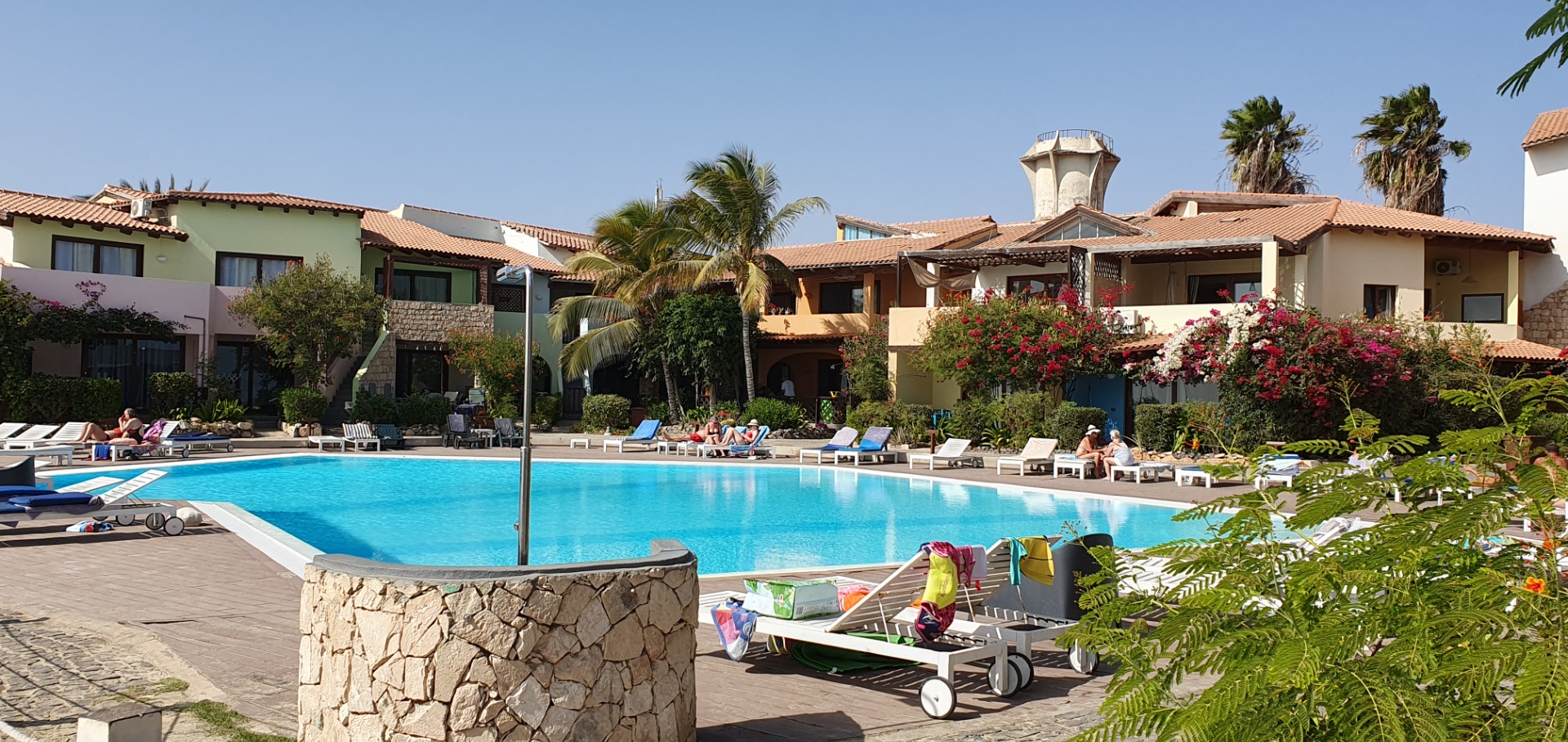 Sunny resort courtyard with a large blue swimming pool, lounge chairs, palm trees, and flowered buildings.