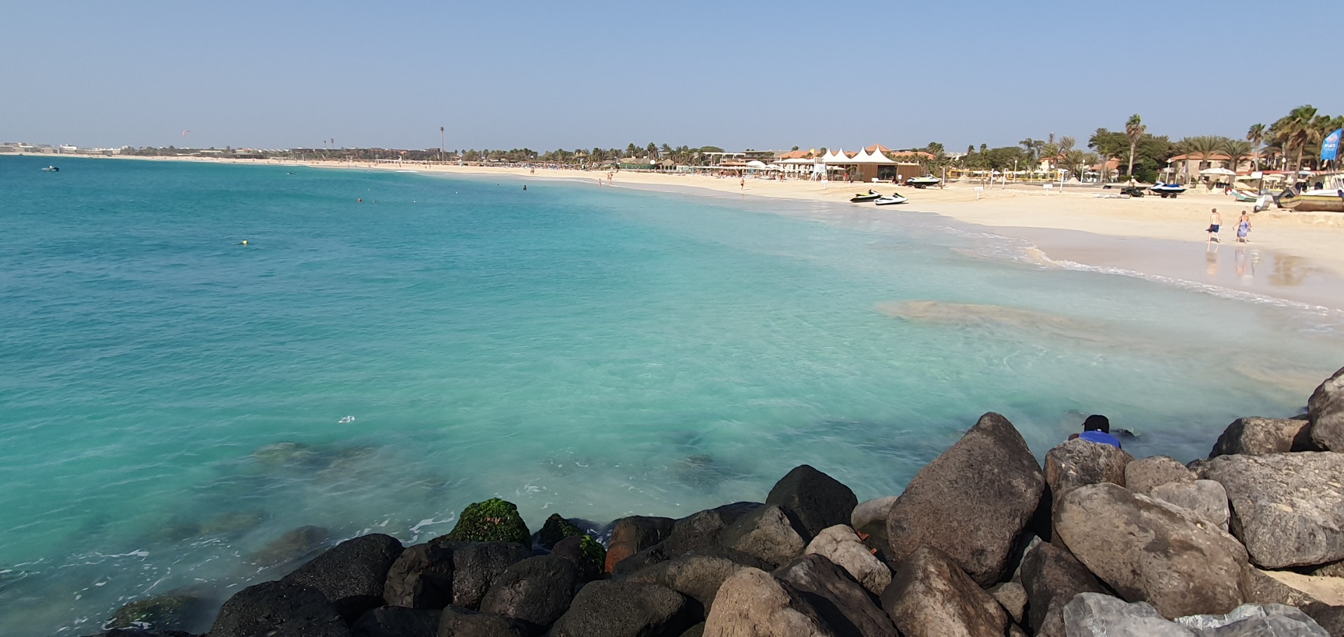 Sal Kapverde - Türkisfarbenes Meer, Sandstrand mit Palmen, Sonnenschirmen und Jetskis, Blick von Felsen am Ufer.