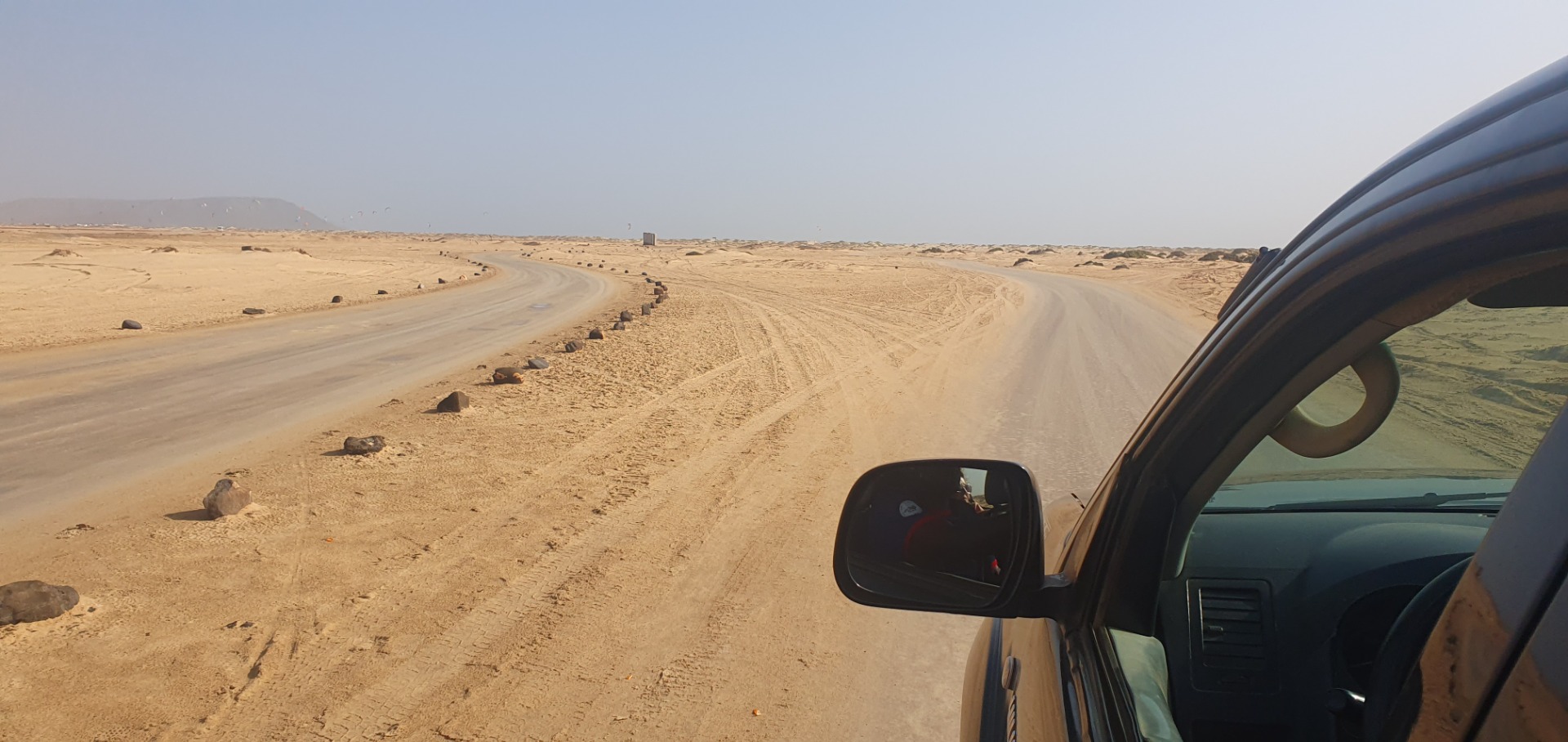 Desert road curves ahead, viewed from inside a car with side mirror and dashboard visible.