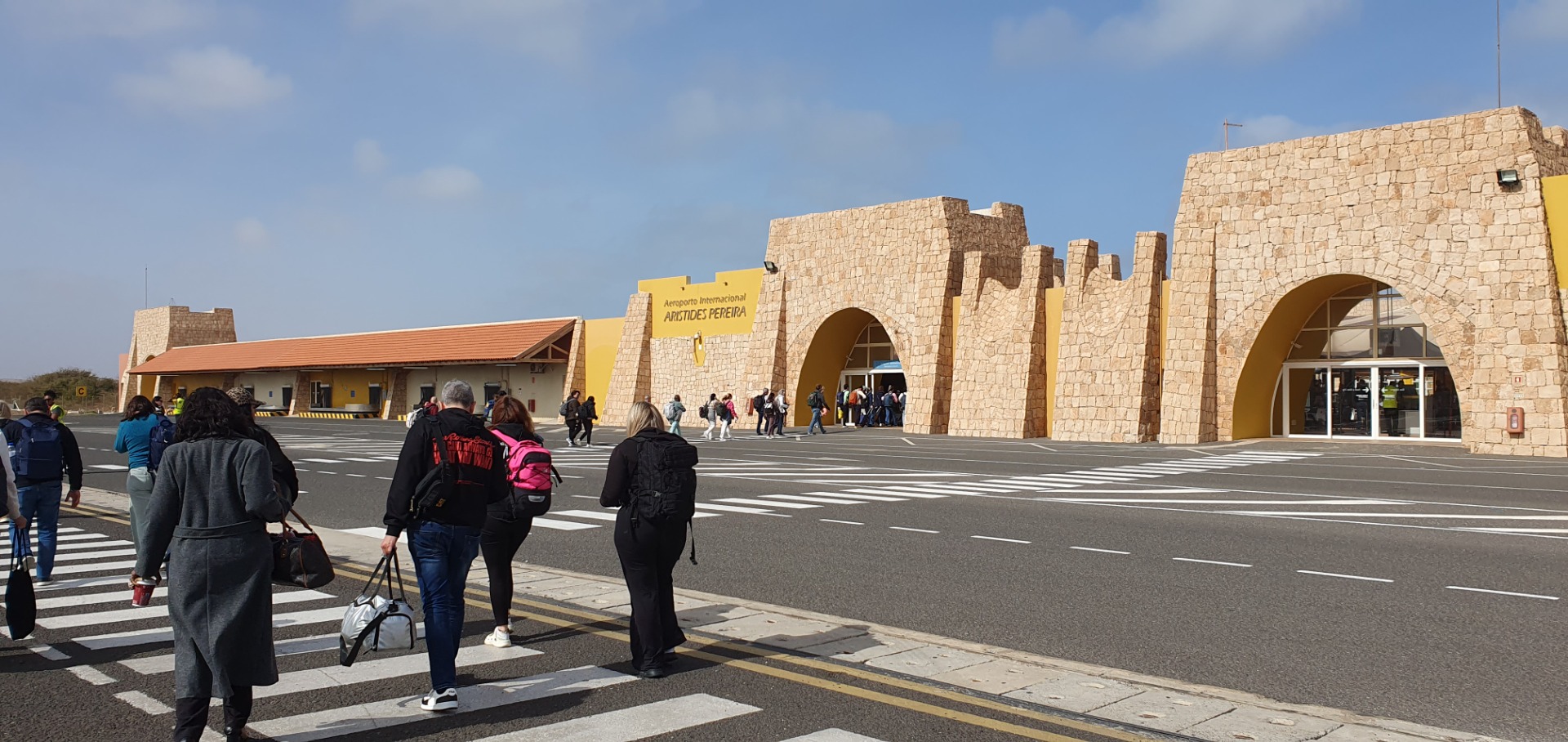 Travelers with backpacks walk toward a stone airport terminal entrance under a clear blue sky.