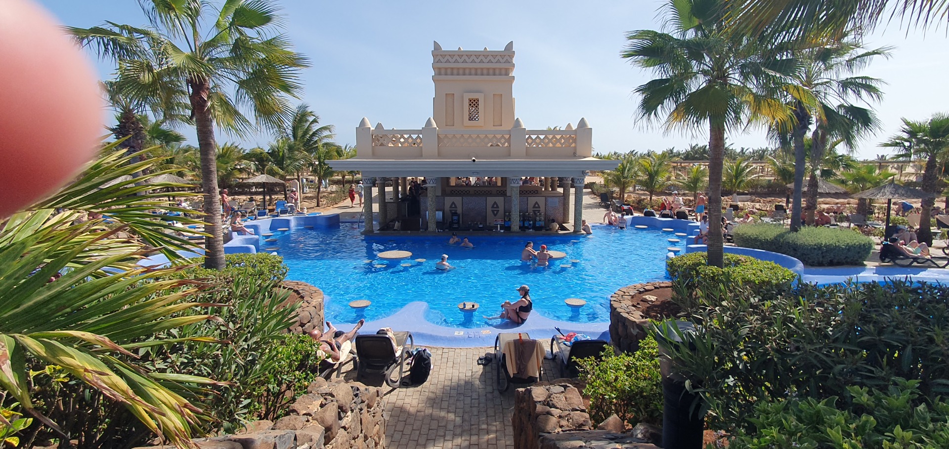 Resort pool with people relaxing, palm trees, and a decorative white tower structure in the background.