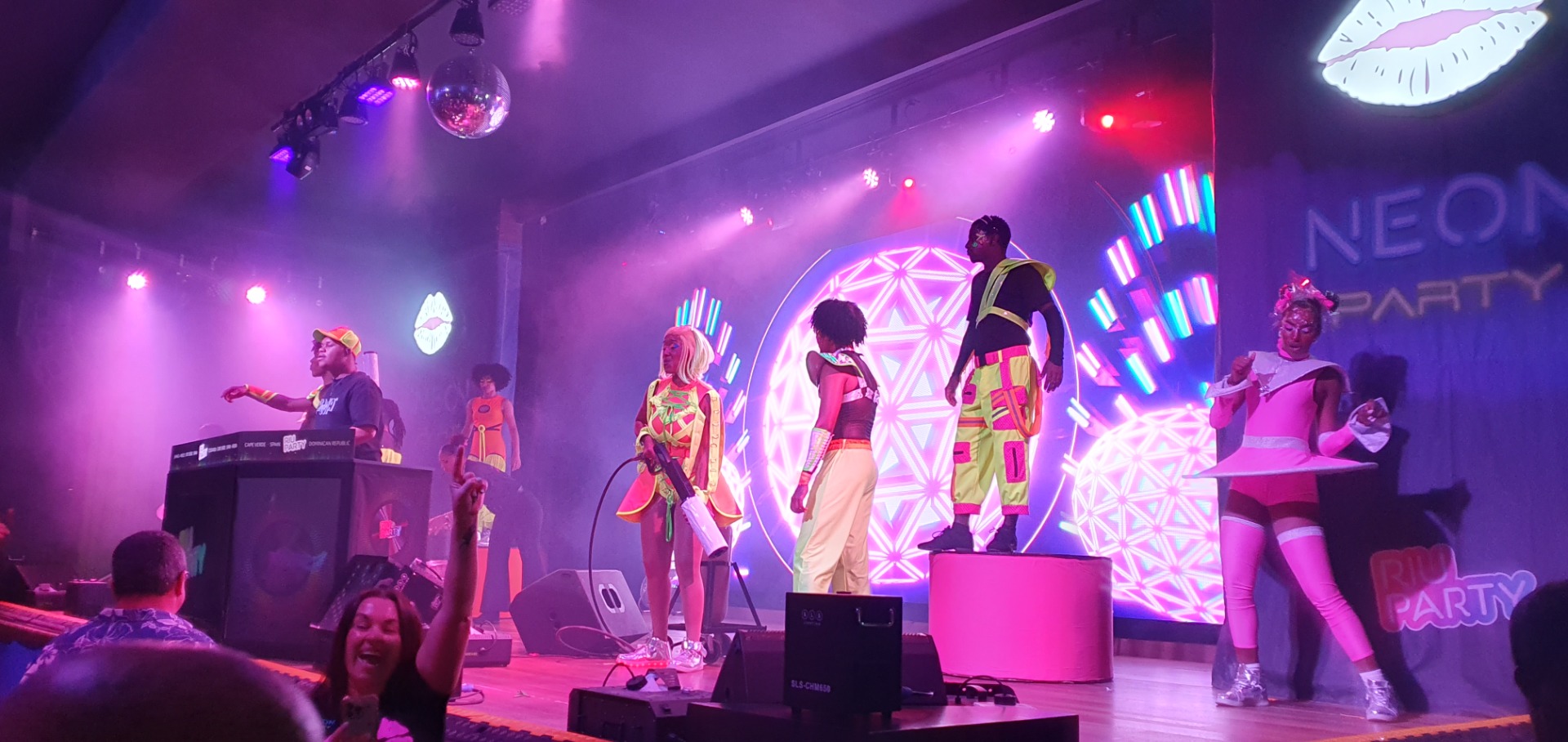 Dancers in colorful outfits perform on a lit stage with purple spotlights and LED backdrop.