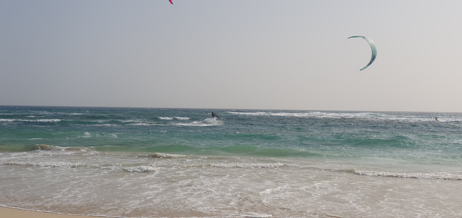 Kite surfer rides choppy green waves on a windy beach, with another kite visible in sky.