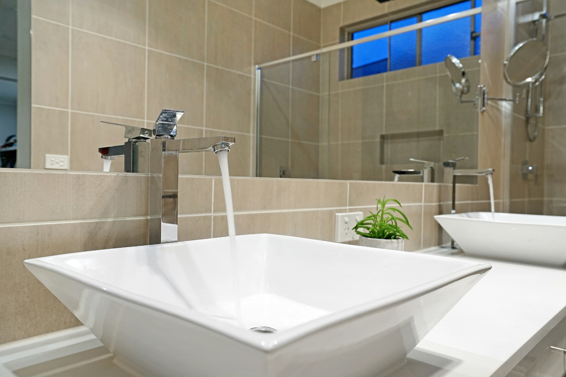 Modern bathroom with white sink, chrome faucet, tiled walls, and glass shower enclosure with blue window.