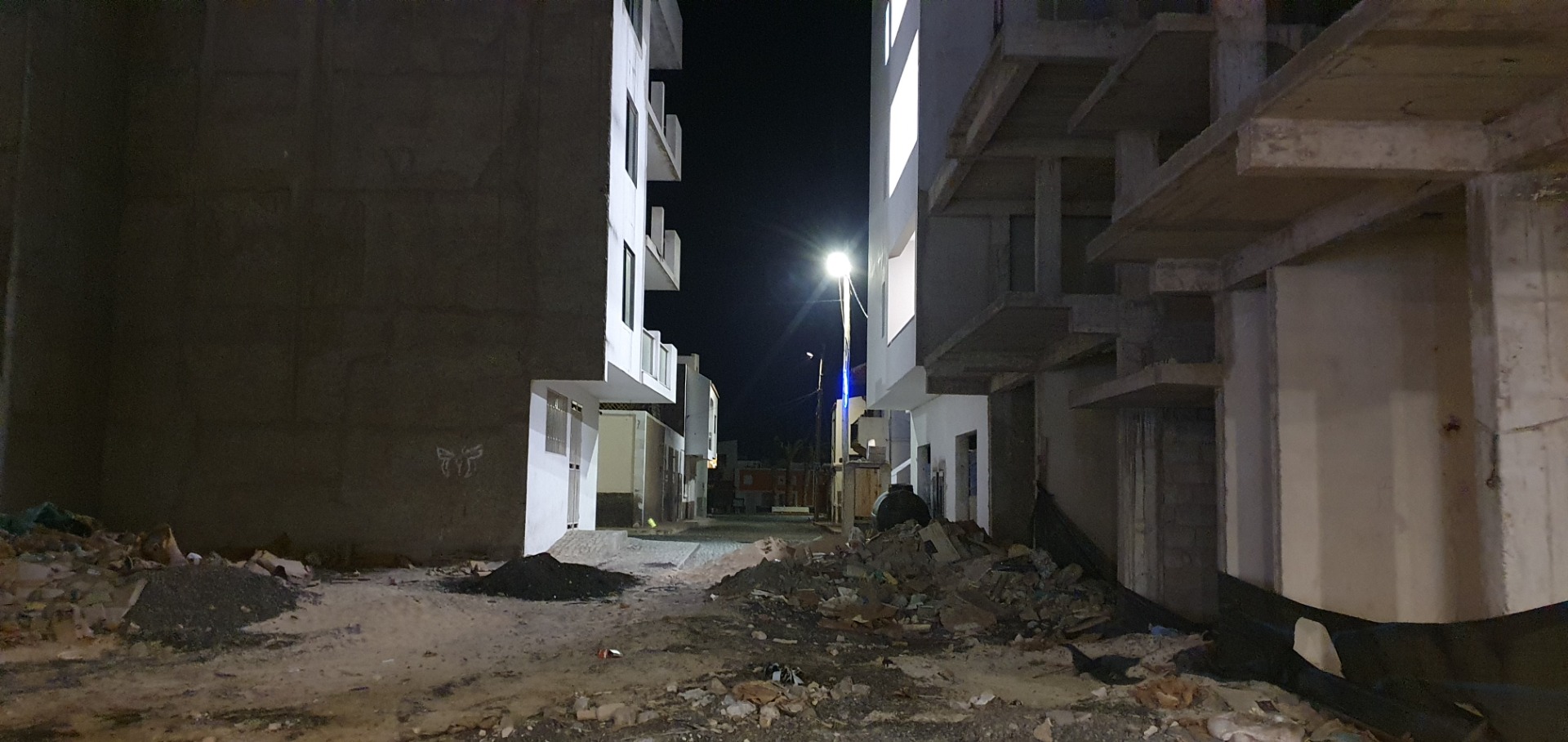 Nighttime view of an unfinished courtyard between white buildings, rubble and debris under bright streetlight.