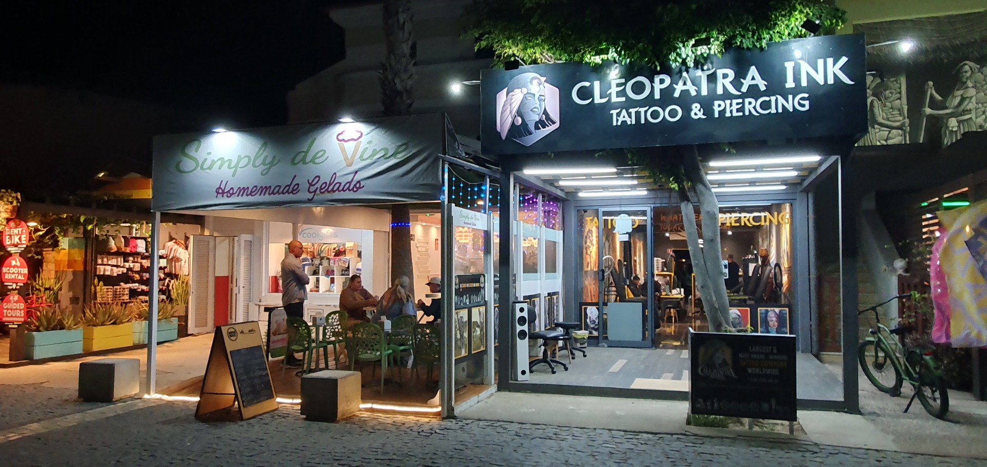 Nighttime storefront with signs for Cleopattra Ink tattoo and piercing, plus Simply de Vive gelato.