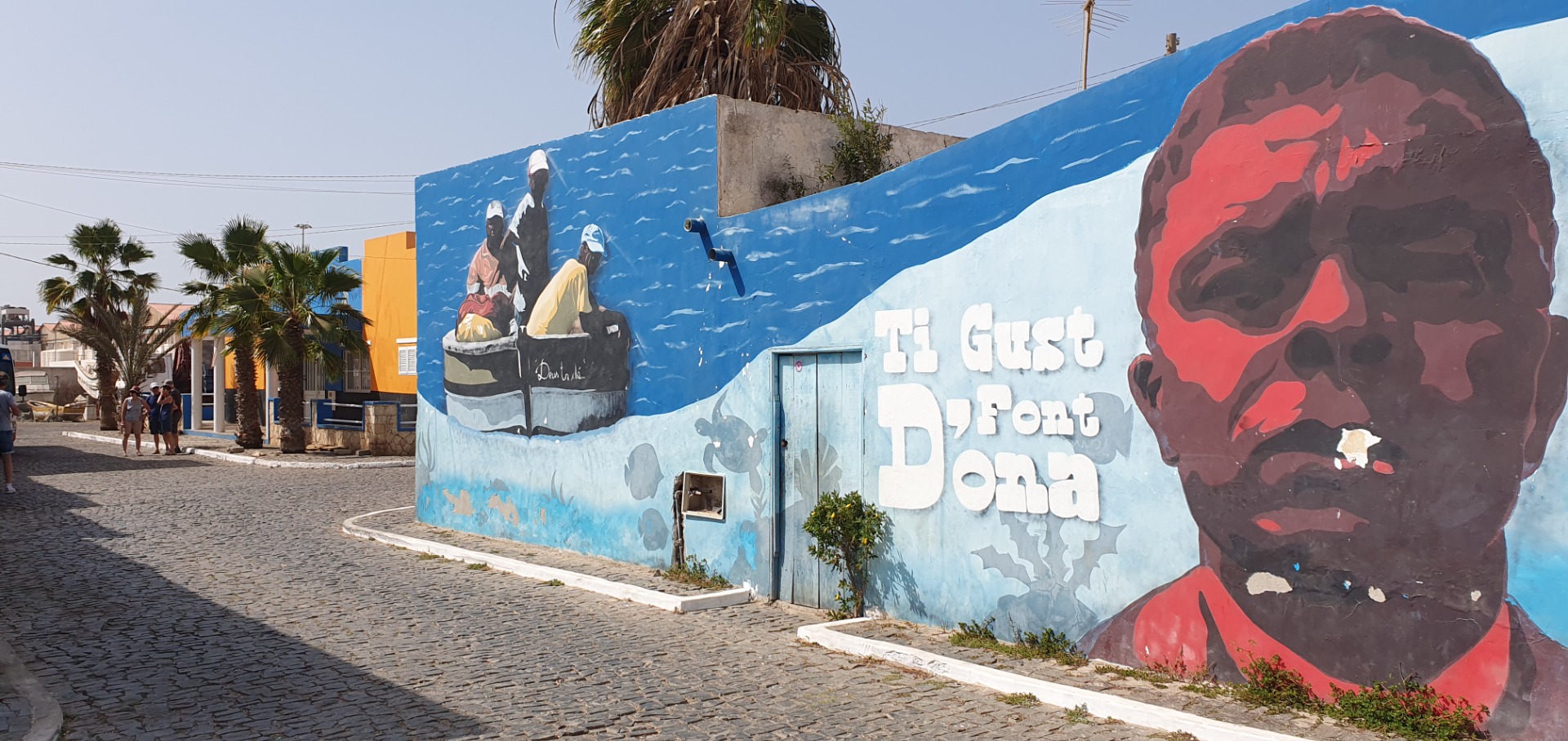 Colorful mural on blue wall with “Hi Guest” text, large red face, palm trees, and cobblestone street.