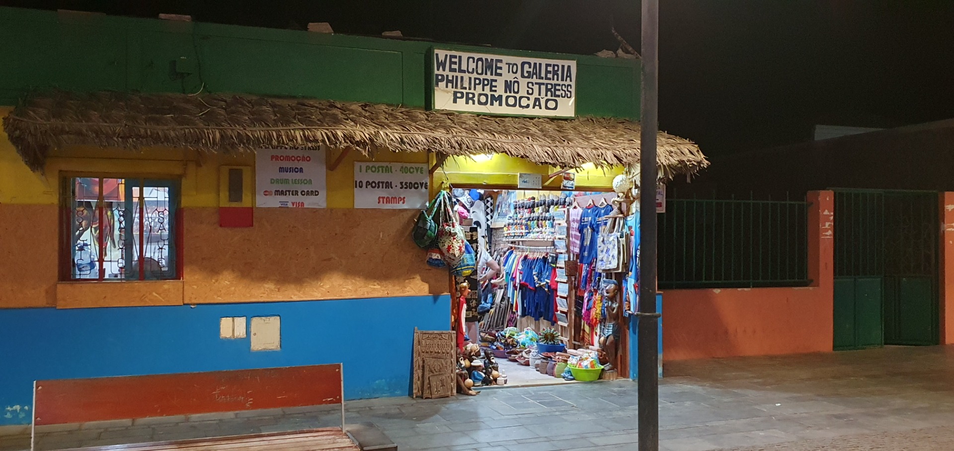 Nighttime storefront entrance with thatched awning, souvenir stalls, and a sign reading “Welcome to Galeria”.