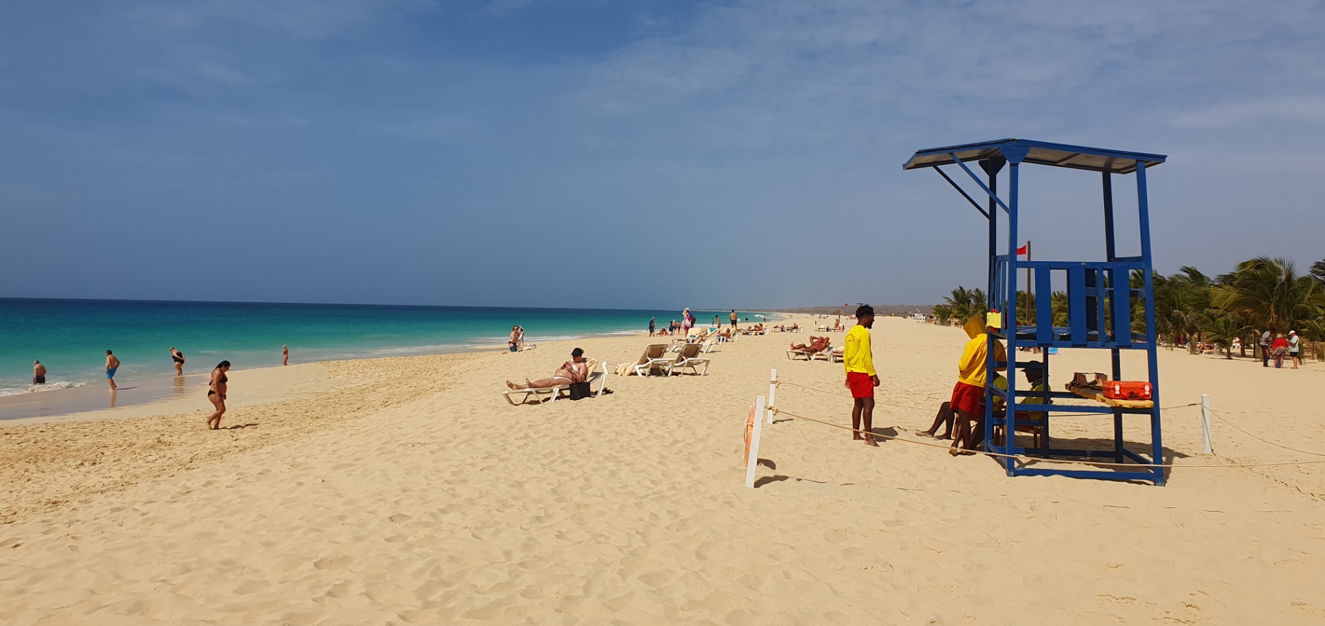 Sunny tropical beach with lifeguard tower, people relaxing on sand, and turquoise ocean under blue sky.
