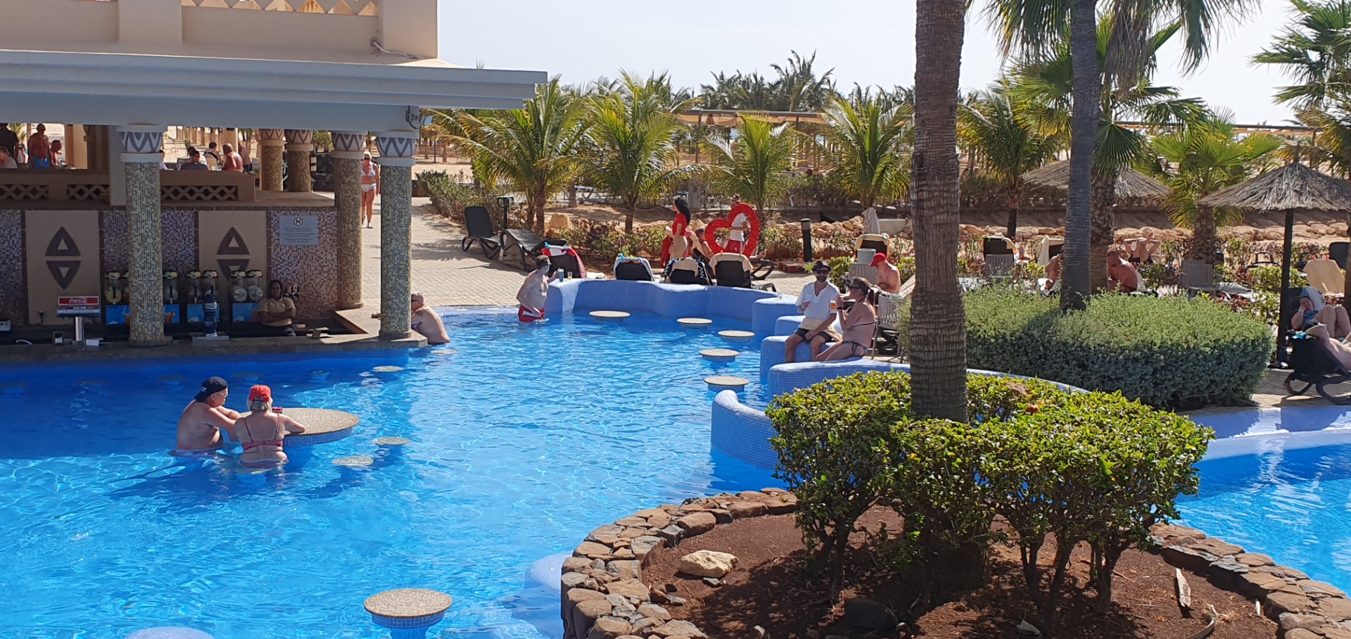 Sunny resort pool with people relaxing, palm trees, and a blue water feature in the background.