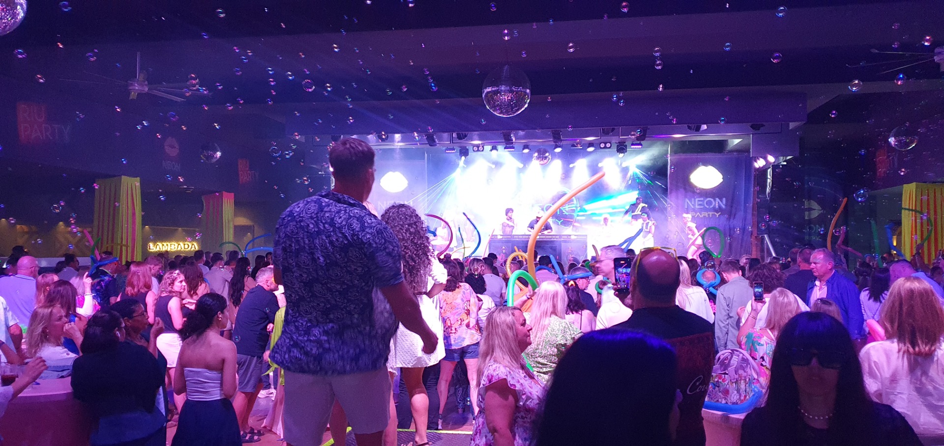Crowd at a neon-themed party, confetti floating above a stage with bright disco lights and performers.