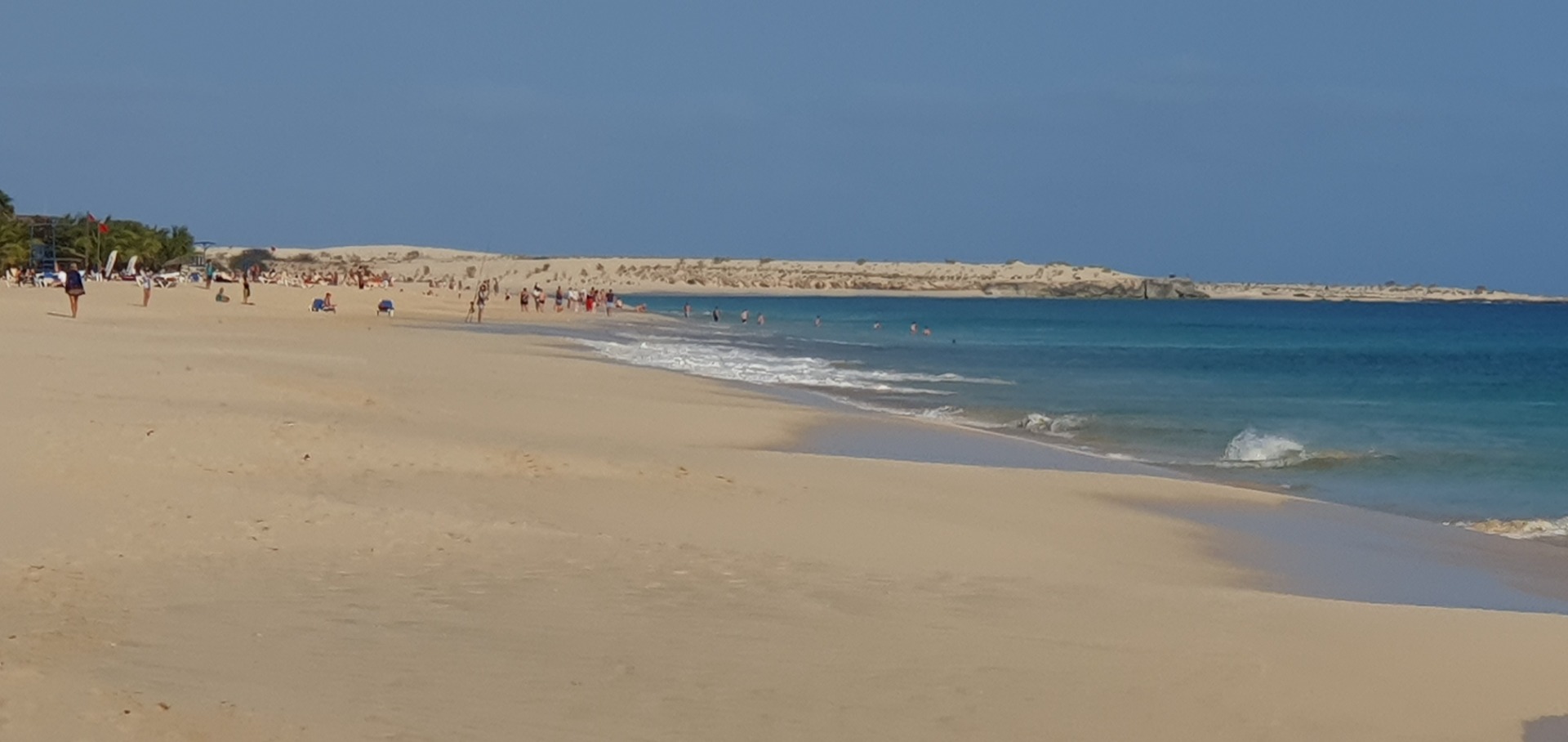 Sunny wide beach with people walking, blue ocean waves, and sand dunes under a clear sky.