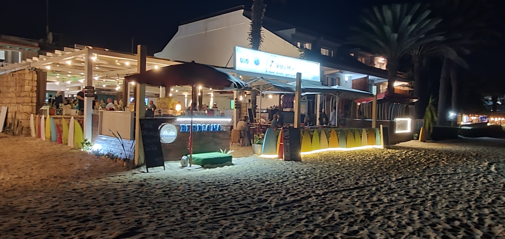 Nighttime beach bar with lit umbrellas, people seated, and a large screen glowing above the counter.