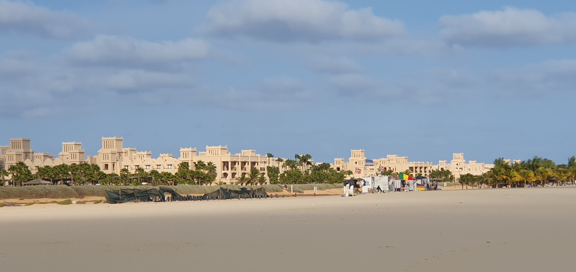 Beachfront resort buildings with palm trees, umbrellas, and sun loungers along sandy shore under cloudy sky.
