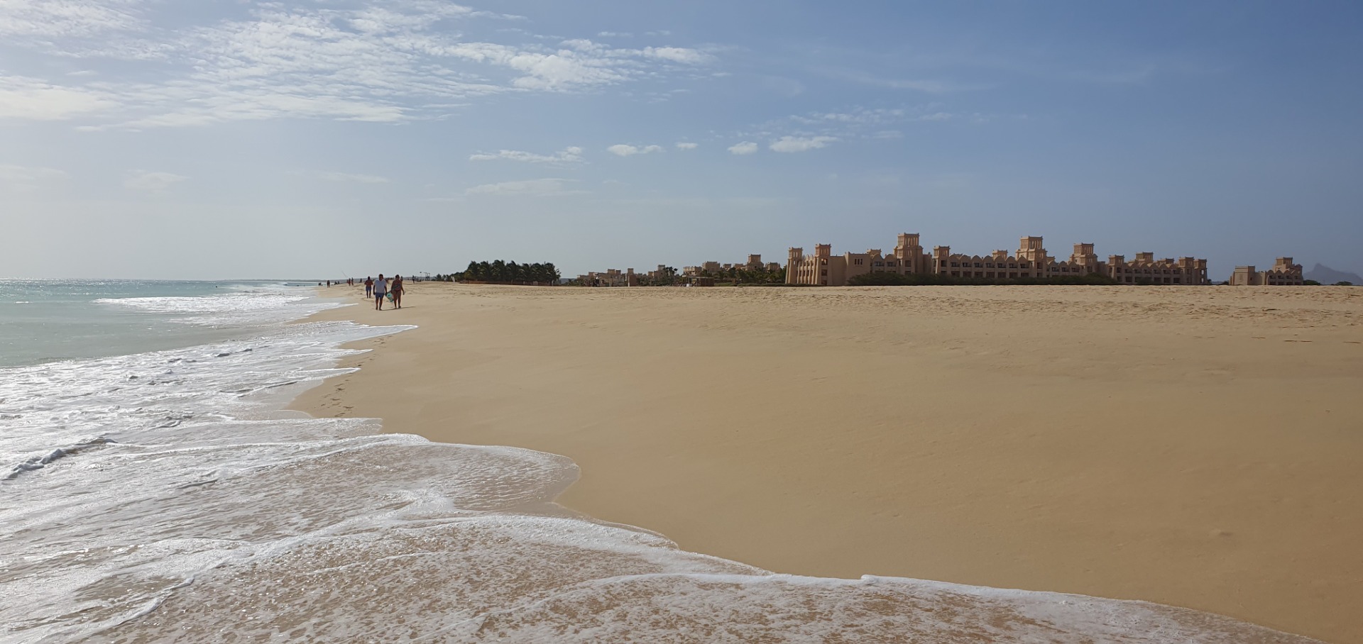 Weicher Sandstrand mit Wellen, Spaziergängern und einer Reihe gelber Gebäude am Horizont unter bewölktem Himmel.