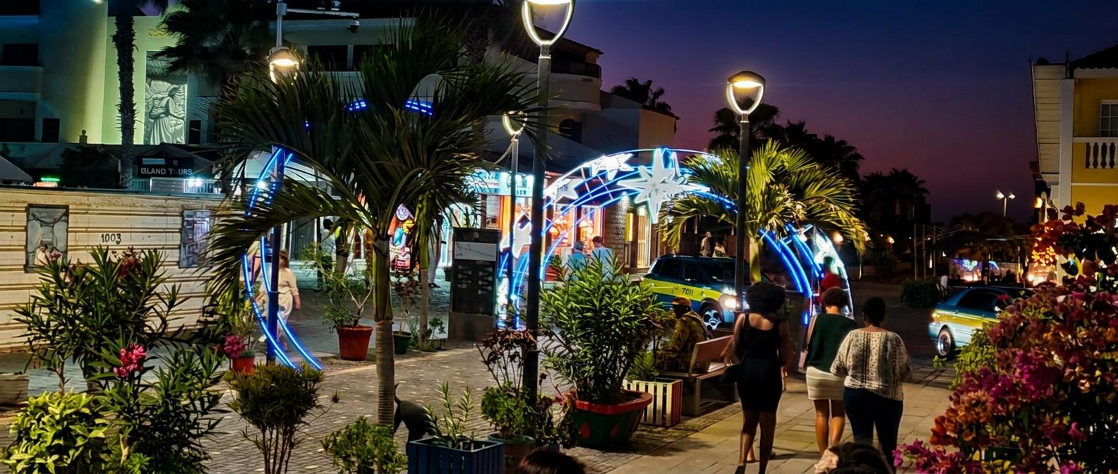 Ilha do Sal - Cabo Verde -Passeio noturno com palmeiras e pessoas, iluminação azul e estrelas em arco, carros e prédios ao fundo.