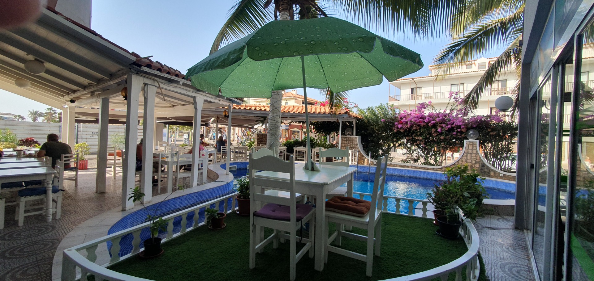 Sal Kap Verde- Terrassenbereich mit weißem Tisch, Stühlen, grünem Sonnenschirm, Pool und Palmen, umgeben von Blumenbeeten.