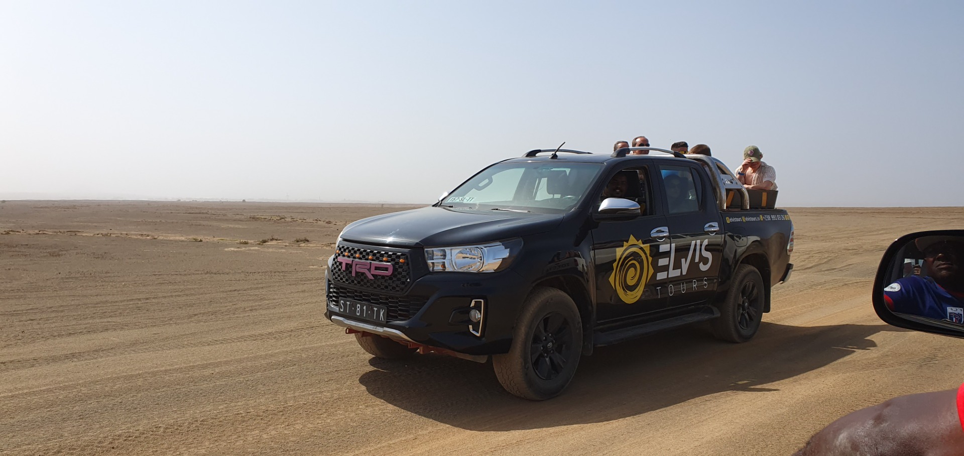 Sal - Cabo Verde- Dunkler Geländewagen mit Aufklebern fährt durch trockene Wüste, mehrere Personen sitzen auf dem Dach.