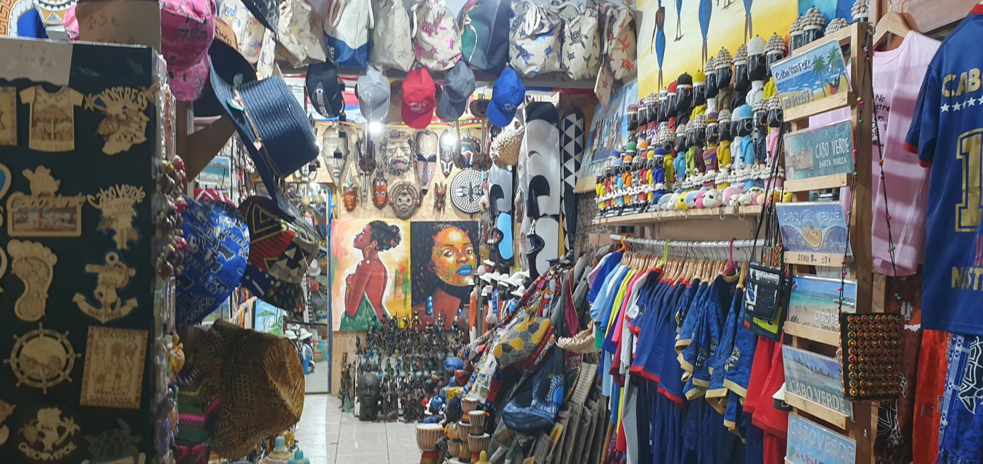 Sal - Cabo Verde - Buntes Souvenirgeschäft mit T-Shirts, Hüten, Masken und Wandbildern in einem engen Marktgang.