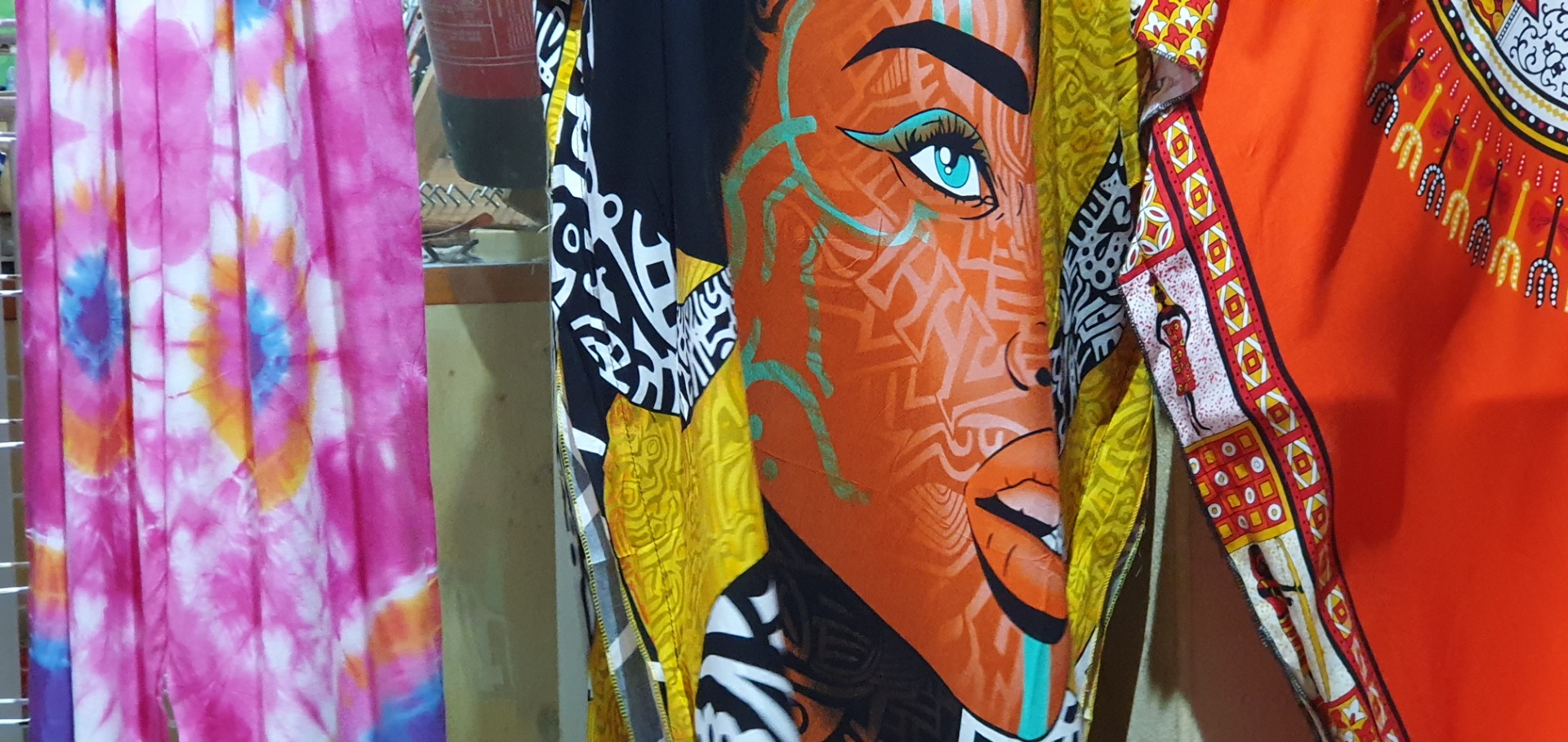 Sal - Cabo Verde- Bunte Graffiti-Gewänder mit stilisierten Gesichtern, orangefarbene Figur mit blauem Auge und pinkem Batikvorhang daneben.