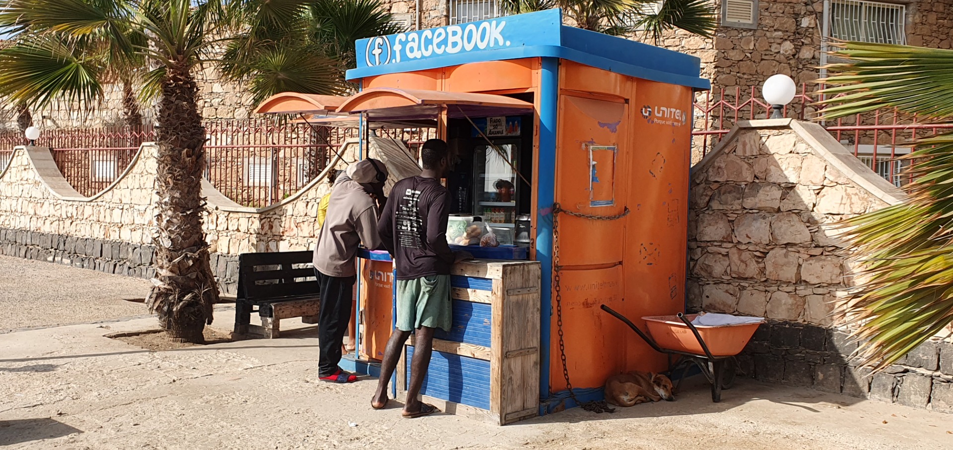 Santa Maria - Sal - Zwei Männer stehen an einem orange-blauen Verkaufsstand mit „Facebook“-Schild, vor Steinmauer und Palmen.