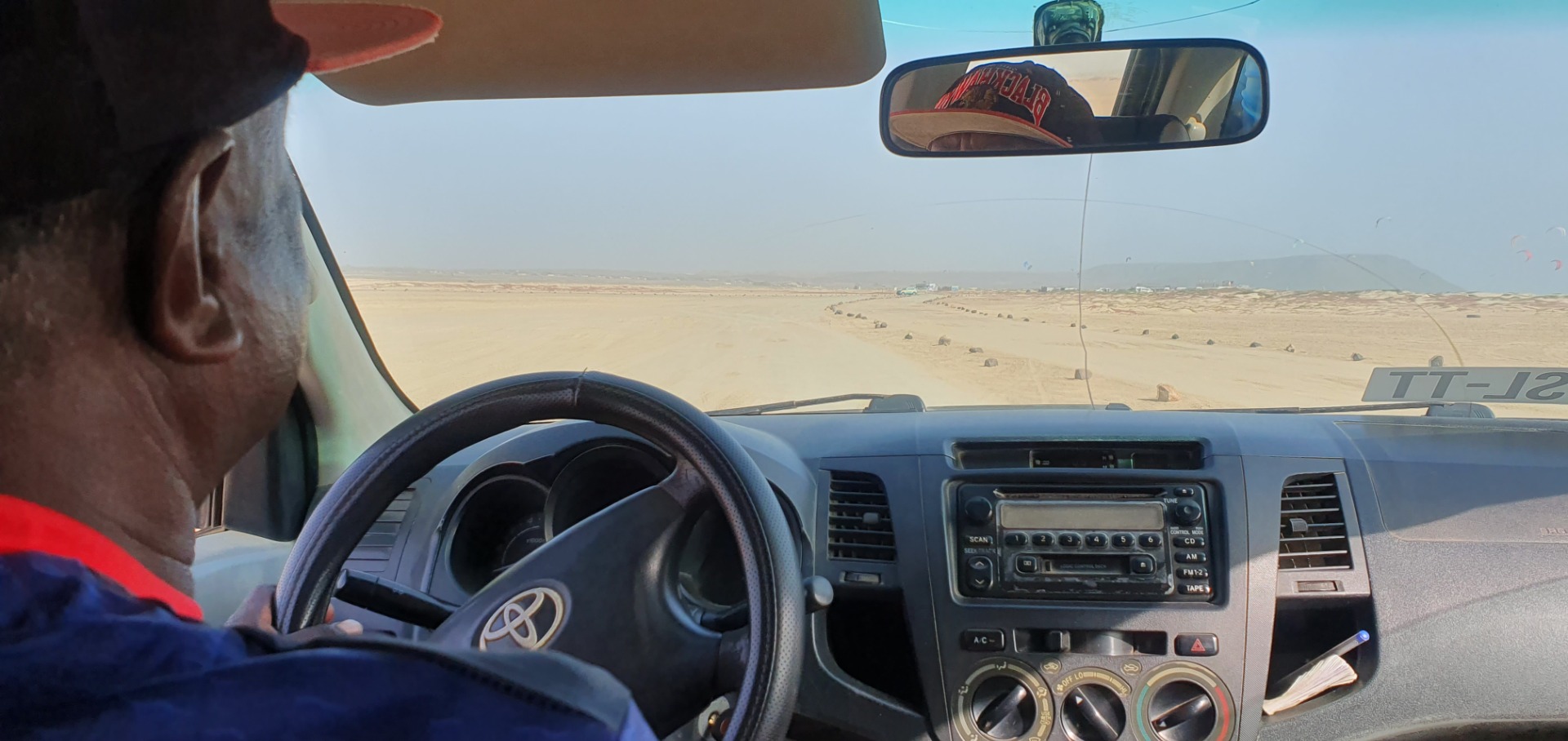 Taxi in Sal - Cabo Verde - Blick aus dem Auto auf Wüste, Fahrerprofil, Lenkrad und Armaturenbrett mit Radio und Klimabedienung.
