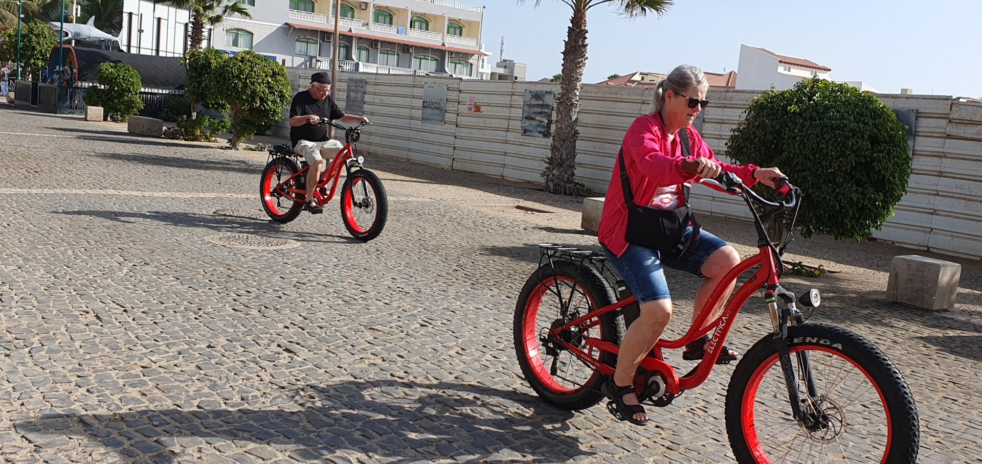 Sal - Cabo Verde - Santa Maria - Zwei Personen fahren auf roten E-Bikes auf gepflasterter Straße, vorbei an Palmen und Hotelgebäuden.