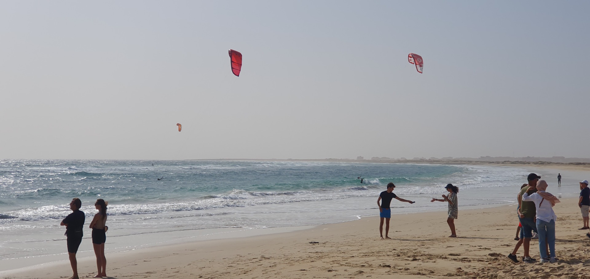 Sal - Cabo verde - Menschen stehen am Strand, während mehrere Kites im Wind über dem Meer schweben.