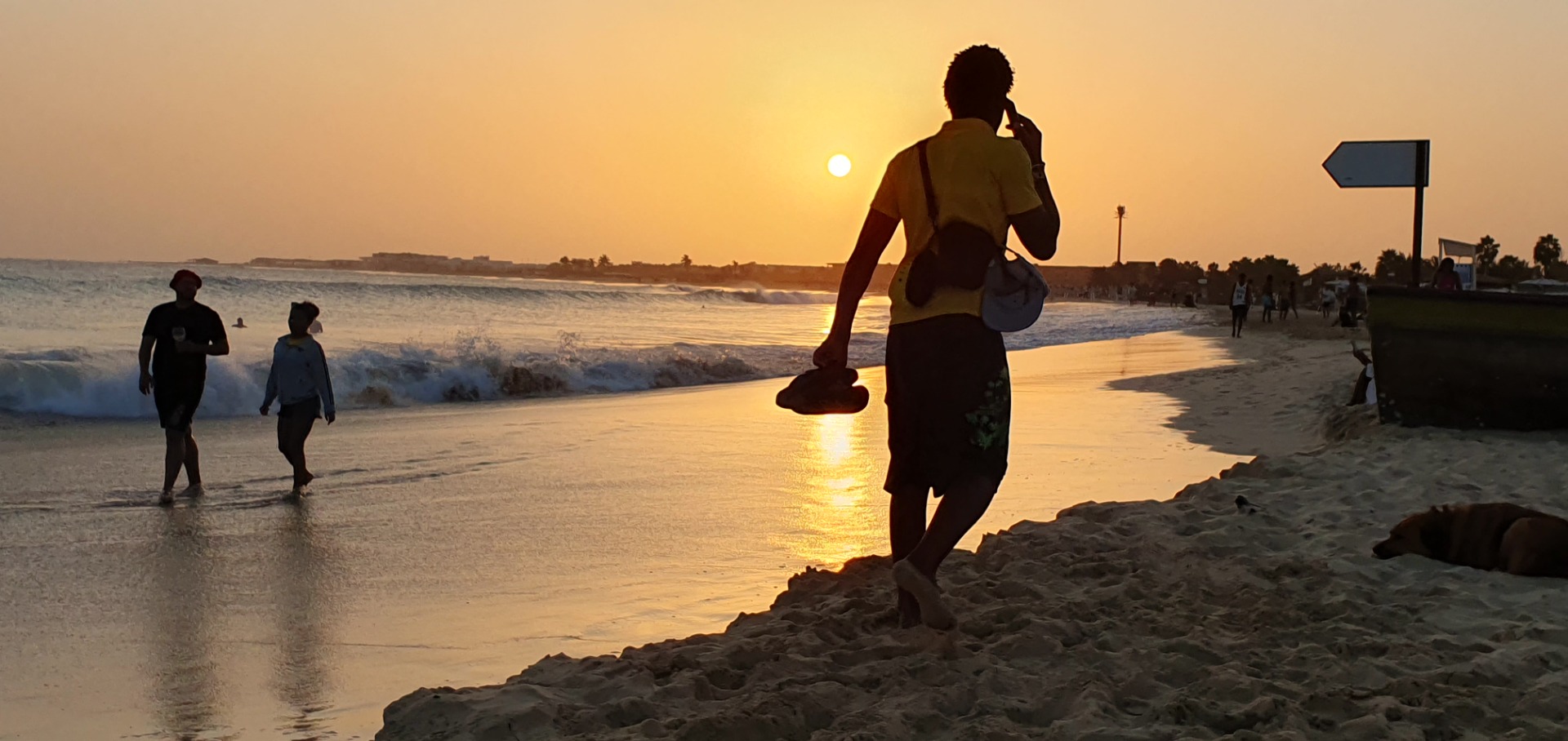 Santa Maria - Sal - Silhouetten von Menschen am Strand beim Sonnenuntergang, eine Person läuft mit Surfbrett, Wellen rollen.
