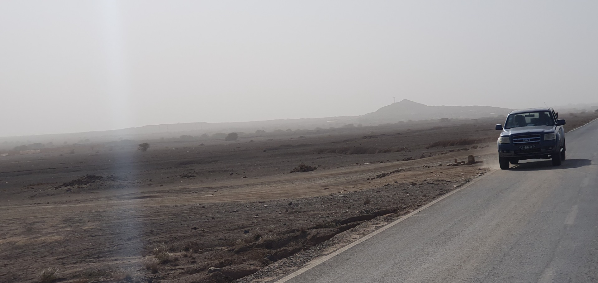 Weißer Lieferwagen fährt auf einer Landstraße durch neblige, trockene Landschaft mit Hügeln im Hintergrund.