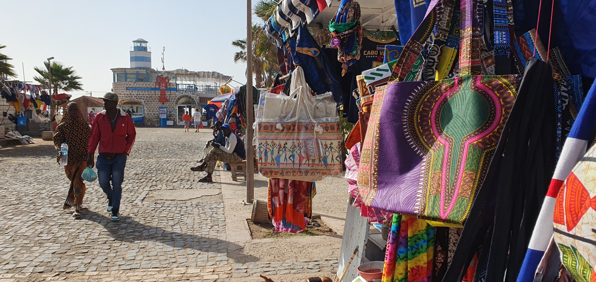 Santa Maria - Sal - Straßenmarkt mit bunten Textilien und Taschen, Menschen gehen auf gepflastertem Platz bei sonnigem Wetter.