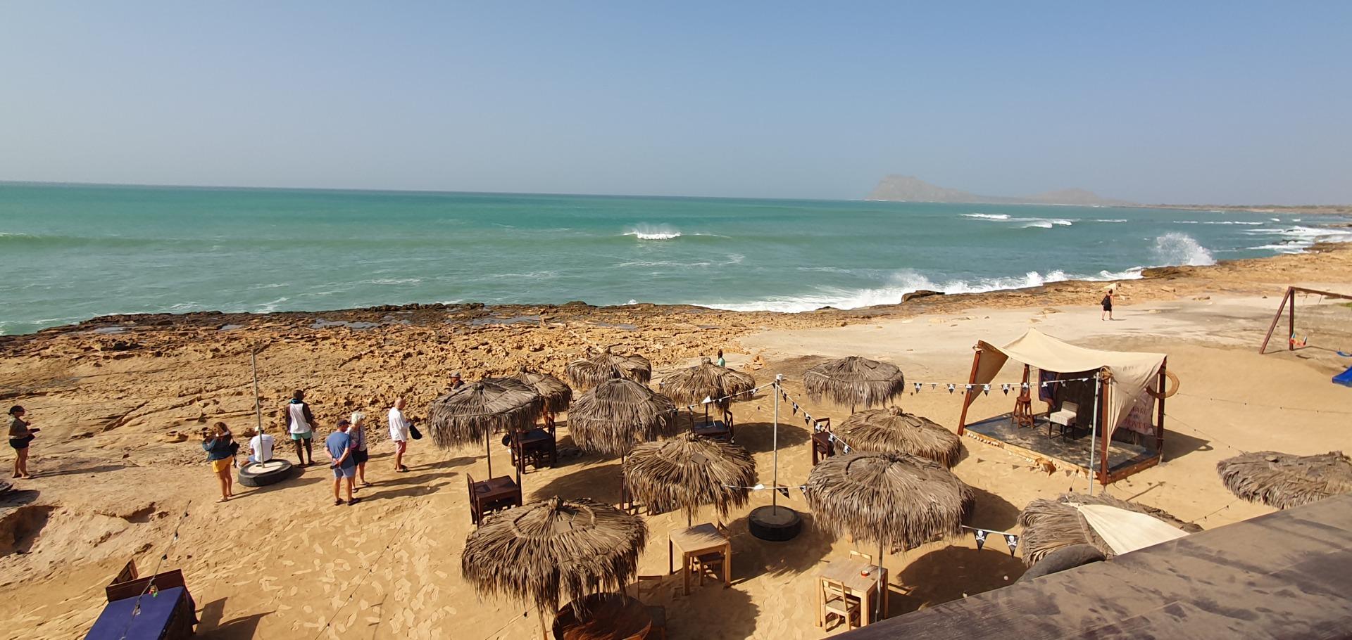 Sal - Cabo Verde - Strand mit Palmenhütten, Sonnenschirmen und Menschen, Blick auf türkisfarbenes Meer und Wellen bei klarem Himmel.