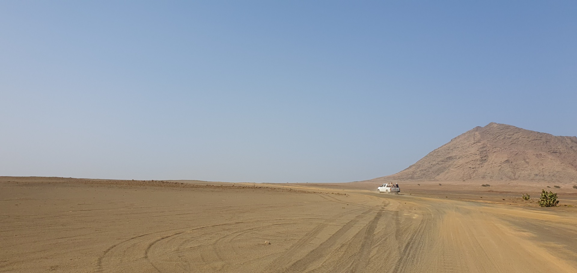 Sal - Cabo Verde -Geländewagen fährt auf staubiger Straße durch trockene Wüste, im Hintergrund ein kahler Hügel unter klarem Himmel.