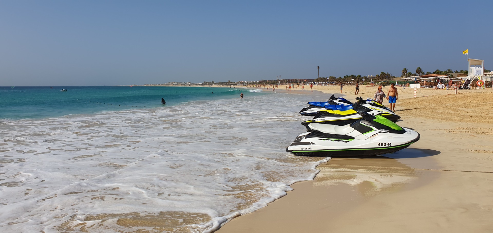 Santa Maria - Sal - Jetski am Strand, türkisfarbenes Meer mit Wellen, Menschen und Strandpromenade im Hintergrund bei klarem Himmel.