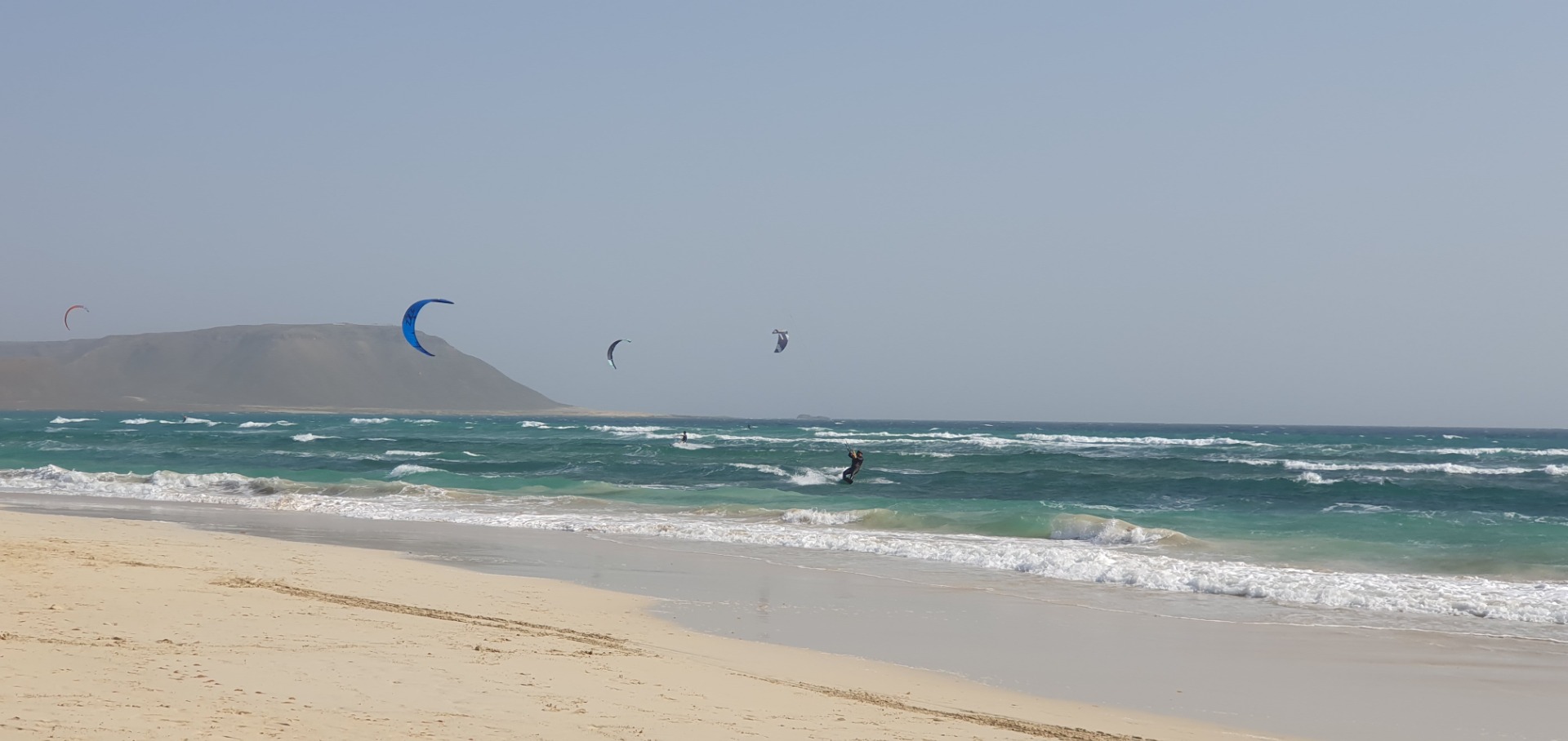 Sal - Cabo Verde - Surfer reiten Wellen am Strand, mehrere Kites im Himmel, windige Küste mit Hügel im Hintergrund.
