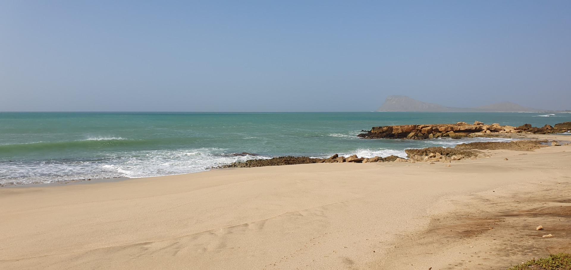 Murdeira - Sal - Cabo Verde - Weicher Sandstrand mit türkisfarbenem Meer, Felsen am Ufer und einer Insel im Hintergrund bei klarem Himmel.