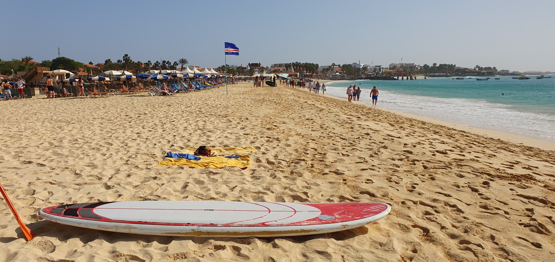 Praia ensolarada com guarda-sóis, pessoas caminhando, bandeira ao fundo e prancha de surf na areia.Praia in Santa Maria, Ilha do Sal